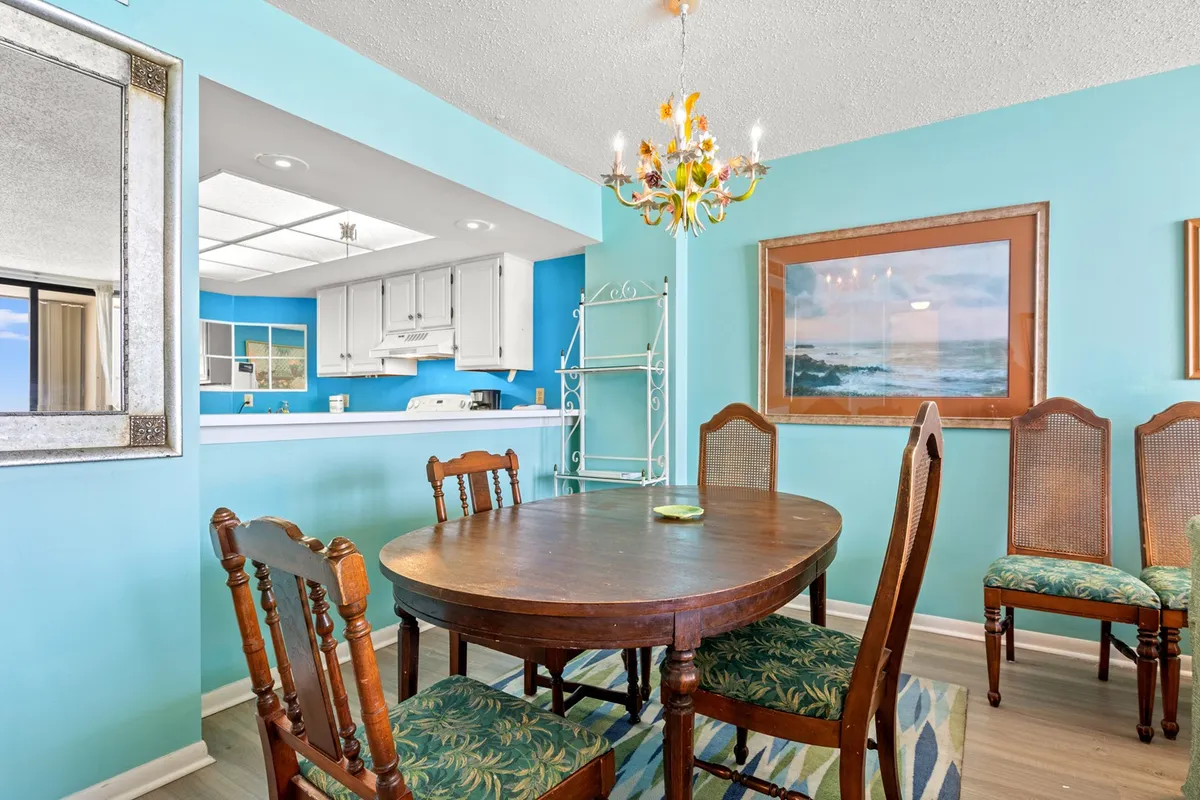 An inviting dining area adjacent to the kitchen, featuring a round wooden dining table under a colorful chandelier, a blue accent wall, and framed beach-themed artwork. The space is designed for family meals, festive gatherings, and easy flow from kitchen to dining.