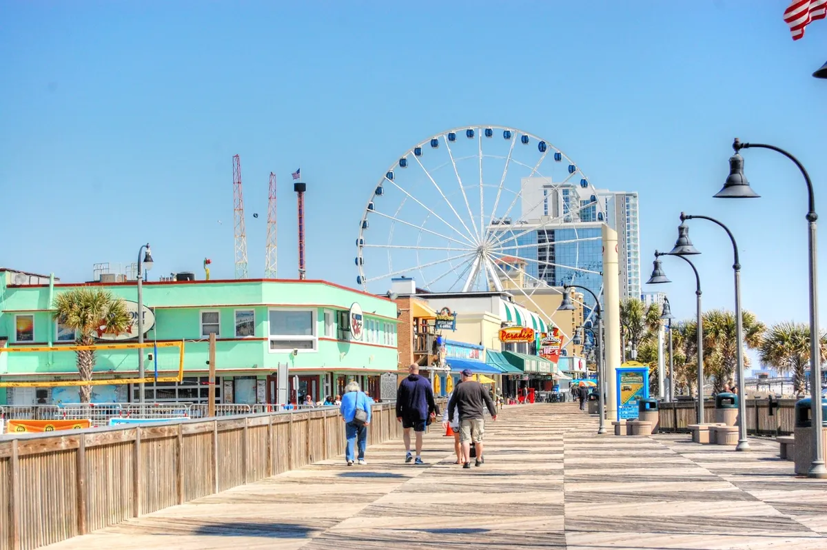 A vibrant boardwalk scene featuring a tall Ferris wheel, colorful buildings, and families strolling along the walkway. The area buzzes with activity, suggesting amusements, rides, and opportunities for family bonding, all set near the beach.