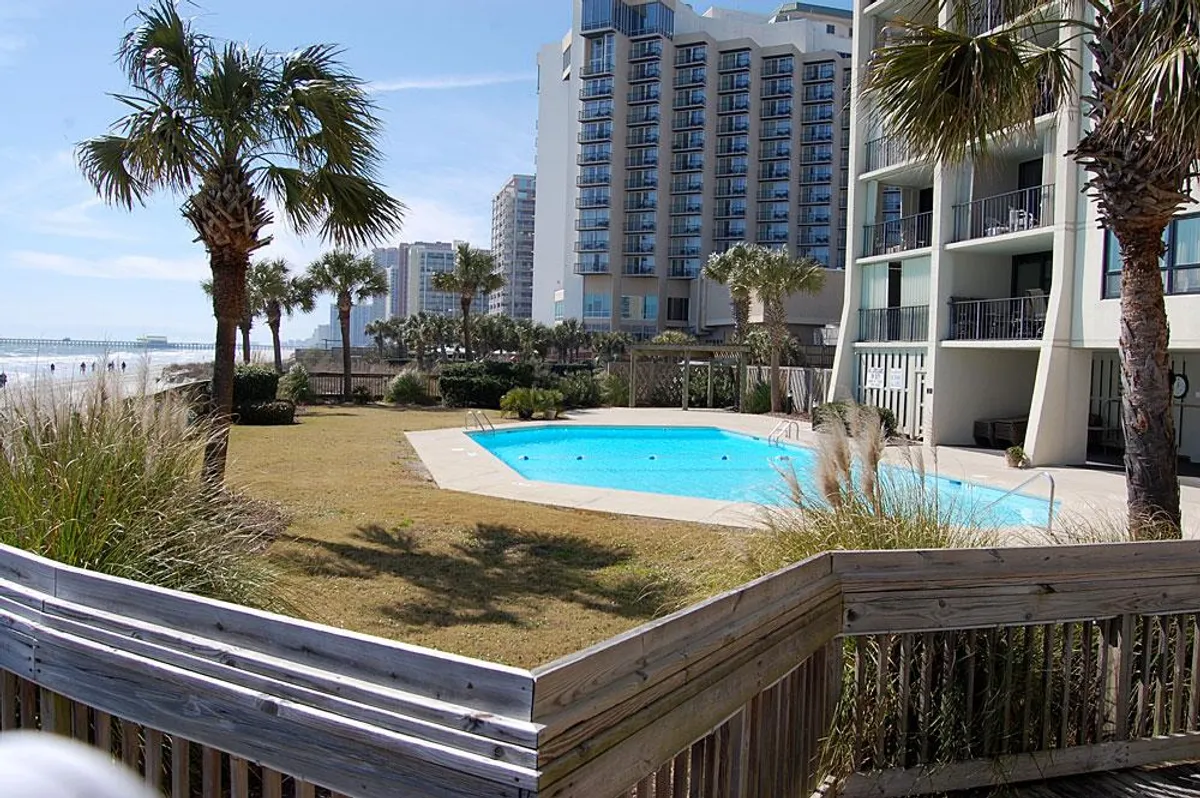 An inviting resort pool framed by palm trees, with a beautiful beach and ocean visible in the background. Guests are seen swimming, sunbathing, and gathering poolside, all set against the tranquil backdrop of the hotel building. The atmosphere is both relaxing and lively, perfect for families seeking a mix of poolside and beachfront activities.