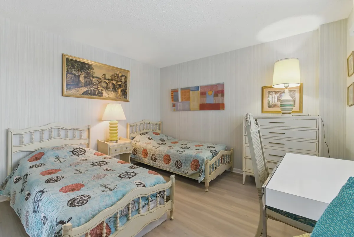 A cozy twin bedroom with two beds adorned in nautical-themed bedding, a bedside lamp casting a warm glow, a wall-mounted painting, and both a dresser and a writing desk. Sunlight streams in, creating a bright and welcoming vibe suited for kids or friends.