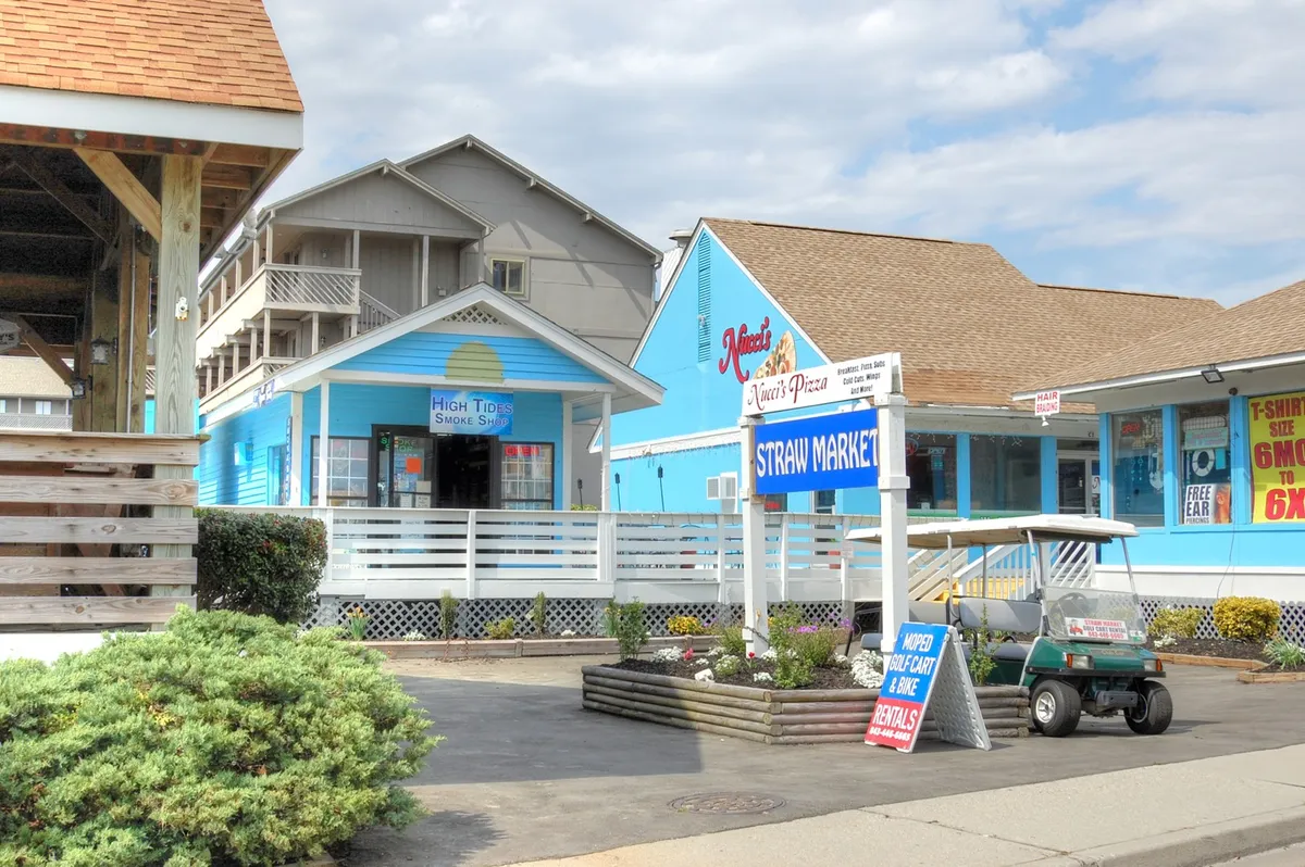 A bustling cluster of colorful buildings and signage for local businesses lines a landscaped area. Visitors walk through the vibrant space, suggesting opportunities for shopping, dining, and exploring local flair together.