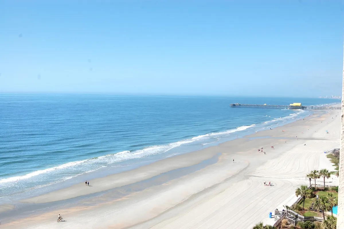 A tranquil stretch of sandy beach hugs calm ocean waves, with a distant pier visible on the horizon. People stroll along the water's edge while a bicyclist enjoys the path, evoking relaxation and a sense of shared adventure.