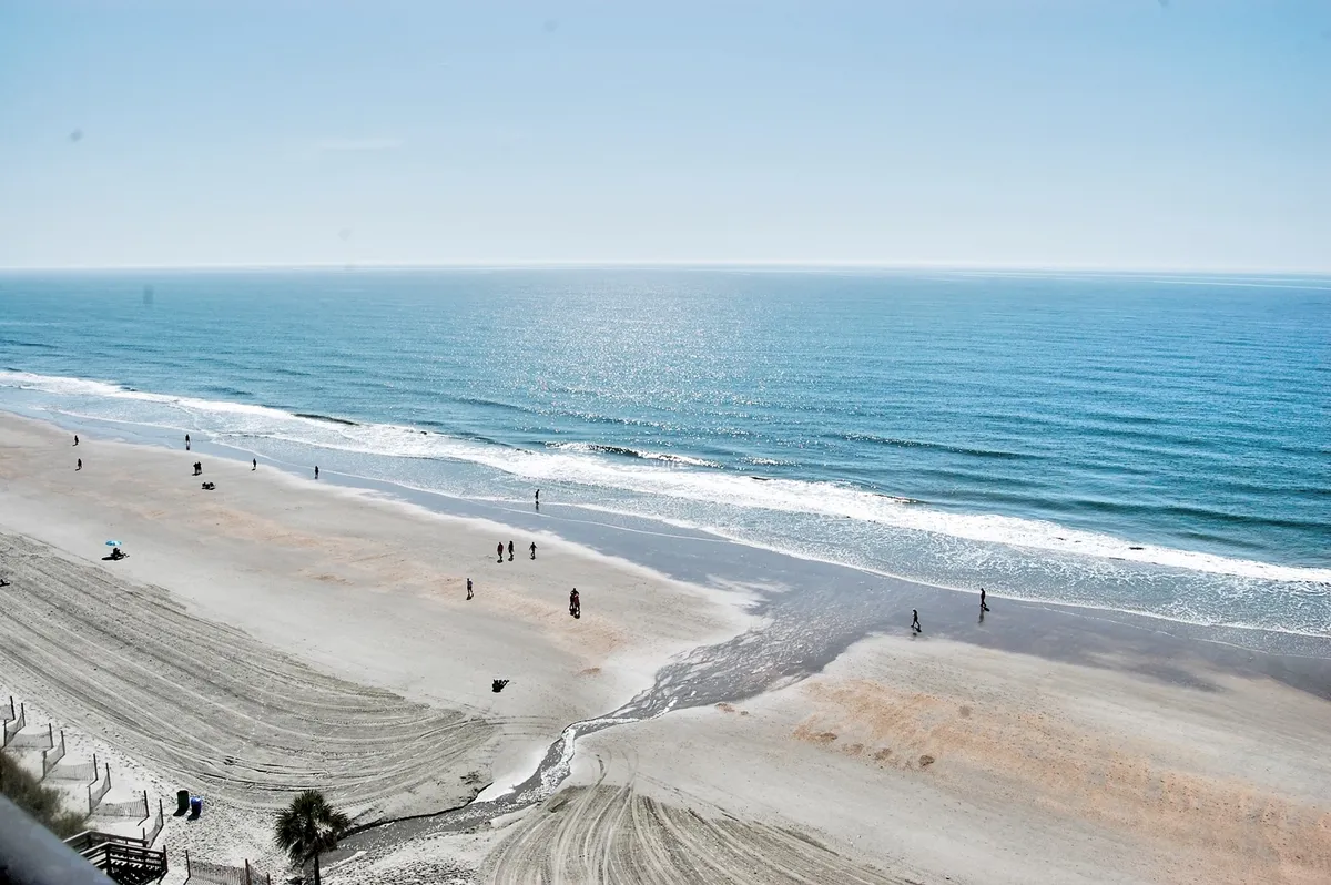 A scenic view of the ocean with gentle waves rolling onto the sandy shore. Families and beachgoers can be seen walking along the shoreline, sunbathing, and playing in the sand—some are likely building sandcastles or preparing for a swim. The scene exudes a relaxed, lively vibe that captures the essence of oceanfront family fun.