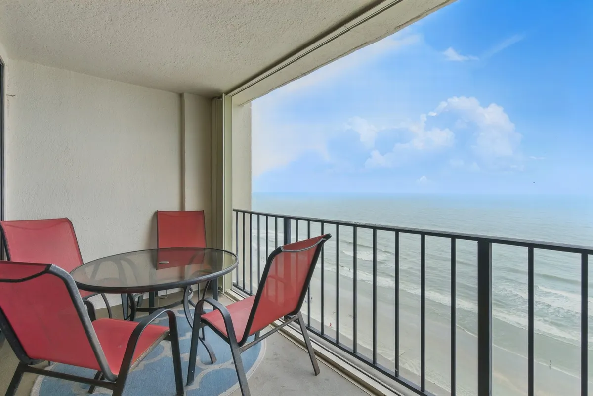 An inviting balcony scene with a glass table, red chairs, and a stunning ocean view, ideal for enjoying breakfast, drinks, or simply soaking in the coastal atmosphere. The proximity to the beach is on full display.