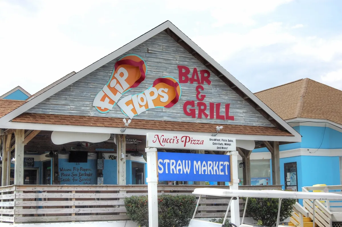 This image captures a lively corner of the local neighborhood, showcasing signs for Flip Flops Bar & Grill, Nucci's Pizza, and the Straw Market. The atmosphere is casual and relaxed, hinting at local dining, shopping, and cultural experiences available to snowbirds during their stay.