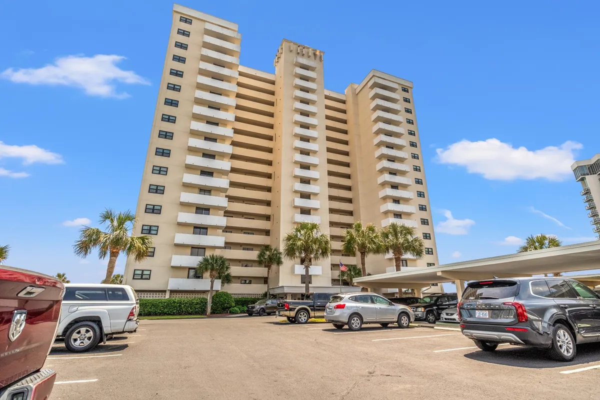 This image displays a tall condominium building surrounded by palm trees and a welcoming parking area. The sun-drenched exterior and lush landscaping establish a sense of arrival and comfortable community living, hinting at resort amenities and the inviting atmosphere awaiting long-term guests.