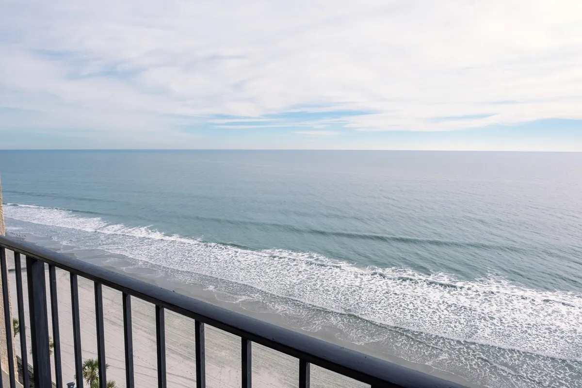 The image features an expansive view from the condo's private balcony, overlooking the endless blue ocean horizon with gentle waves and a textured sky, evoking tranquility. The scene suggests guests relaxing on the balcony, feeling the ocean breeze, and possibly spotting dolphins or watching a stunning sunset.
