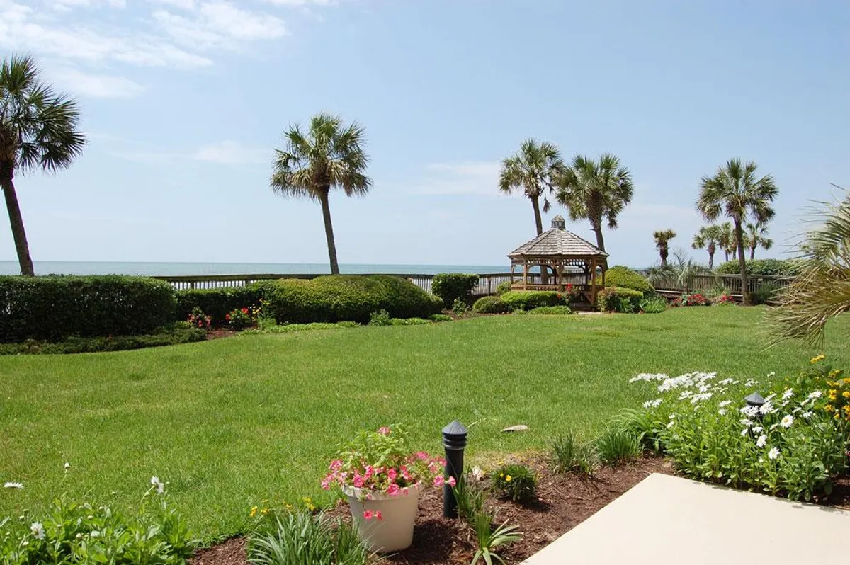 A peaceful exterior scene featuring a gazebo set on a lush green lawn with vibrant flower beds, all overlooking the ocean. The tranquil atmosphere is enhanced by gentle waves in the background and ample space for relaxing or socializing outdoors.
