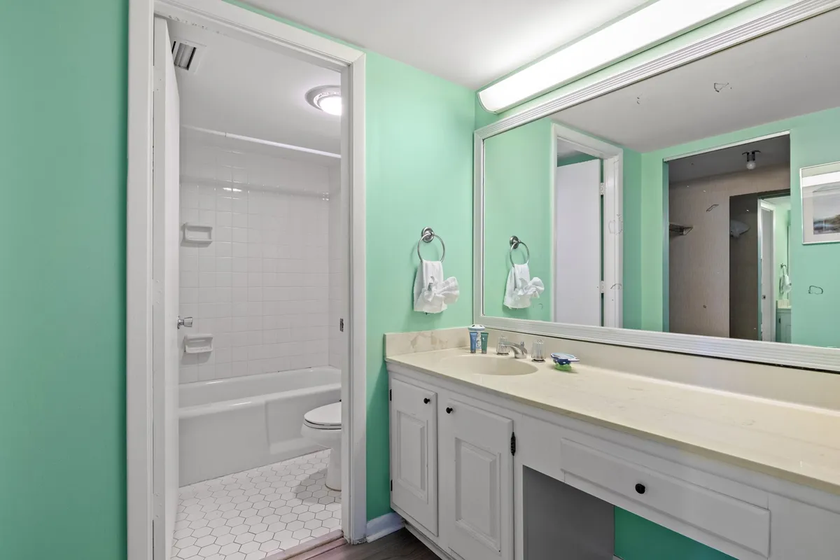 A calming bathroom with mint-green walls, an expansive vanity mirror, white cabinetry, and stylish hexagonal floor tiles. The space is well-lit and feels fresh, inviting, and well-maintained, suggesting a pleasant morning routine or a relaxing evening unwind.