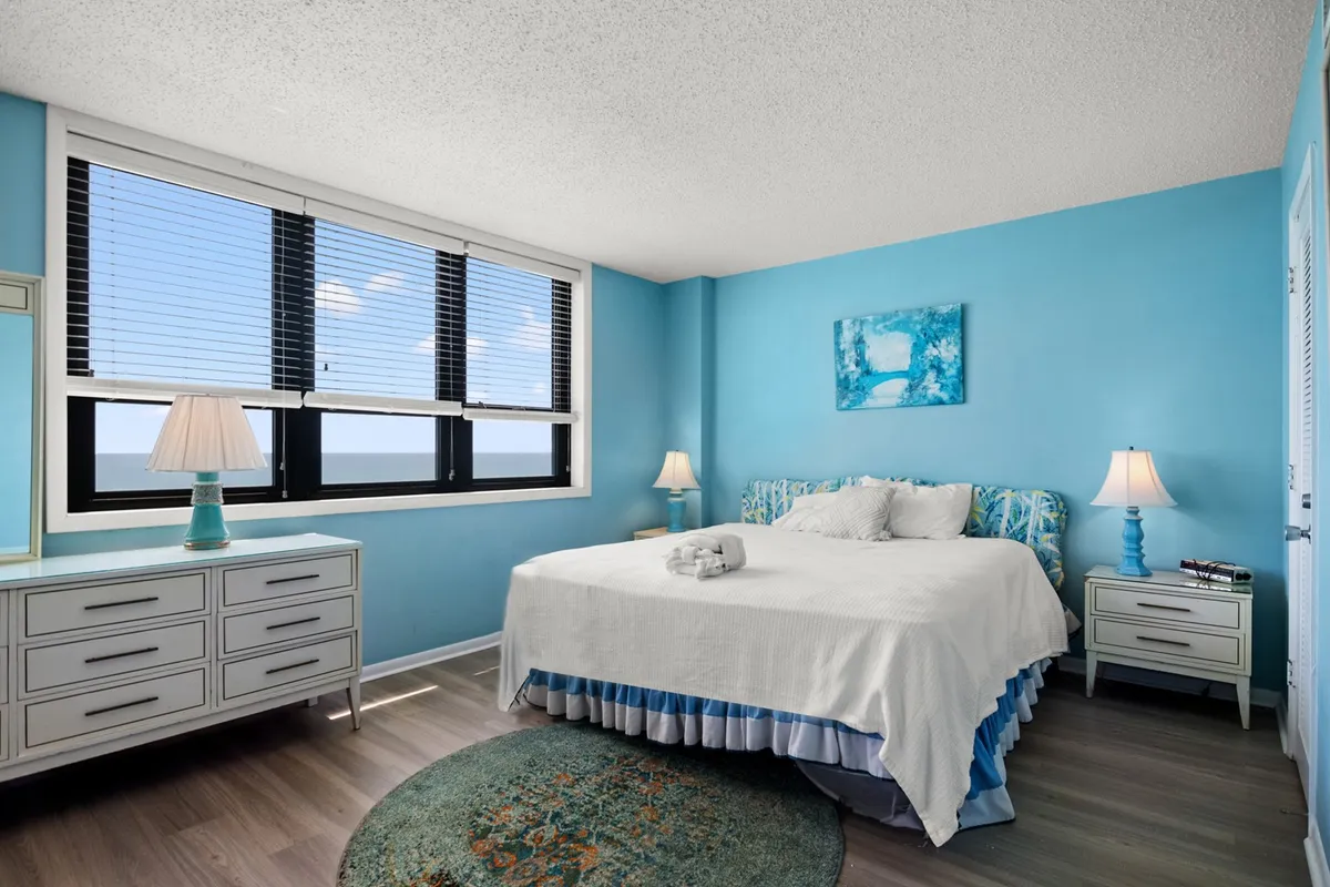 A serene king bedroom is shown with a comfortable bed positioned near a large window revealing an ocean view. The room features a soft area rug, a nightstand with a lamp, a dresser, and gentle natural light that enhances the tranquil mood.